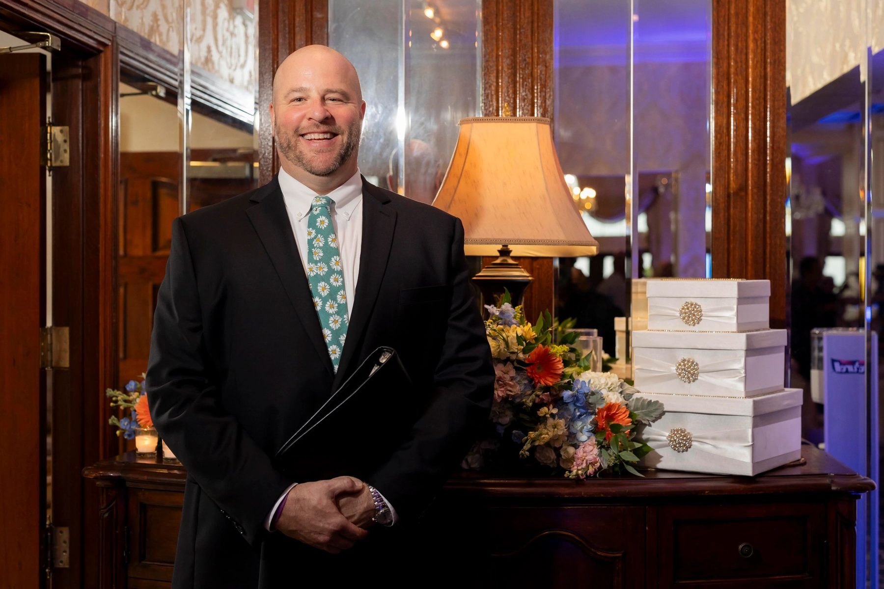 Jim Hill smiling in a dark suit and daisy tie at a wedding venue